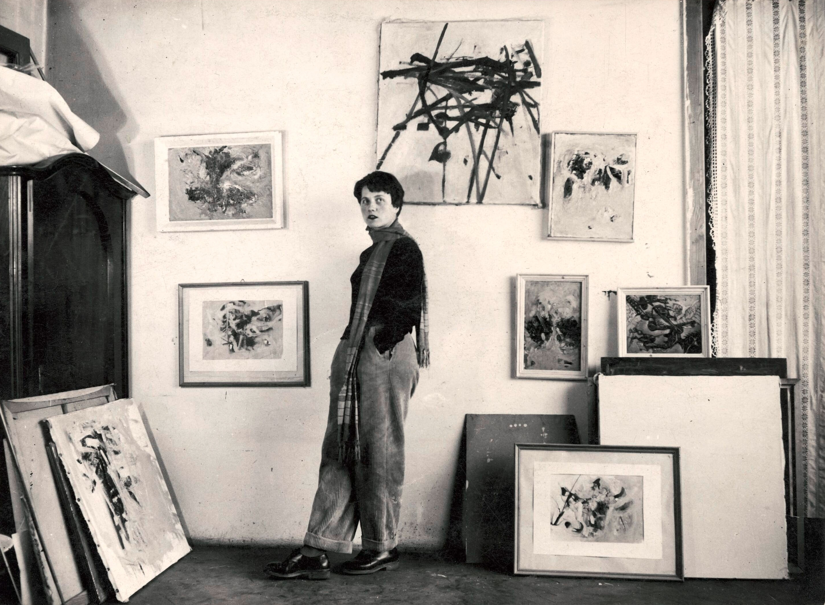 Maria Lassnig, Atelier Heiligengeistplatz (1951) Junge Frau vor einer weißen Wand mit mehreren Gemälden